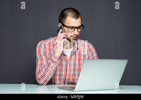 Schöner Mann im Hemd bekleidet und am Telefon sprechen, während mit Laptop über Grau Stockfoto