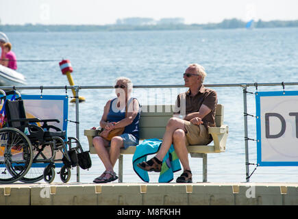 Älteres Paar entspannen Sie an der Wasserlinie auf einer Bank sitzen mit dem Rollstuhl für Neben der Bank gesperrt, am Haringvliet See in Hllevoetsuis Stockfoto