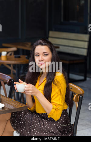 Frau mit langen dunklen Haaren, tragen gelbe T-Shirt, braun Polka Dot Rock und roten Lippenstift trinken Cappuccino in einem Café mit dunklen Innenraum. Vert Stockfoto