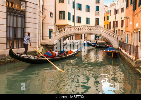 Bunte Gondel Fahrt mit Touristen und Apartments mit Rollläden mit Blick auf eine elegante weiße steinerne Brücke über einen schmalen Kanal in Venedig Stockfoto