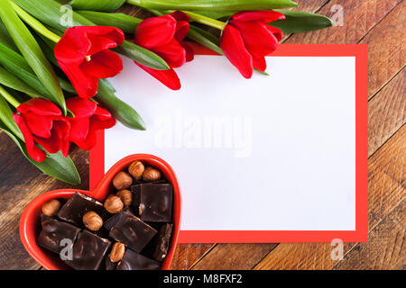 Grußkarte. Weißes leeres Blatt im roten Rahmen mit Kopie Raum, roten Tulpen und rote Herzförmige Schale mit Schokolade Stücke auf rustikalen Holzmöbeln Hintergrund. Stockfoto