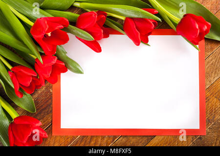 Grußkarte. Weißes leeres Blatt im roten Rahmen mit Kopie Raum und rote Tulpen auf alten rustikalen Holzmöbeln Hintergrund. Stockfoto