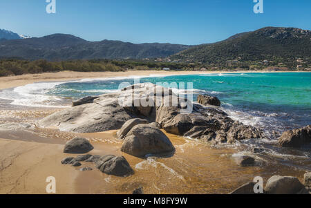 Wellen auf Felsen und Sand am Strand Algajola in der Balagne Korsika unter einem tiefblauen Himmel mit Schnee bedeckte Berge in der Ferne Stockfoto