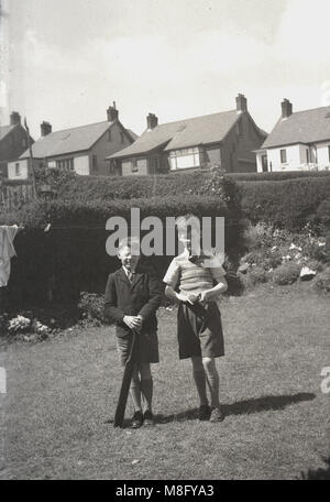 1950, historische, Sommer und zwei lächelnde junge Jungen, vielleicht Brüder, zusammen draußen stehen in einem Vorstädtischen zurück Garten, der Jüngste mit einem alten Kricketschläger, England, UK. Stockfoto