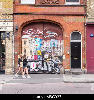 Die Menschen wandern Vor eine alte Tür in Aufkleber und Street Art Wandbilder in Commercial Street, Spitalfields, Shoreditch, East London, UK abgedeckt Stockfoto