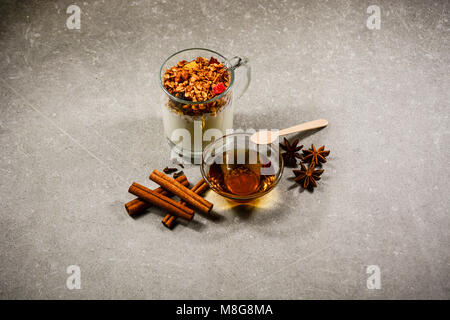 Müsli mit getrockneten Beeren, kandierten Früchten, Nüssen, Joghurt, goldenem Honig, Anis, Zimtstangen und Nelken auf steinernem Hintergrund. Stockfoto