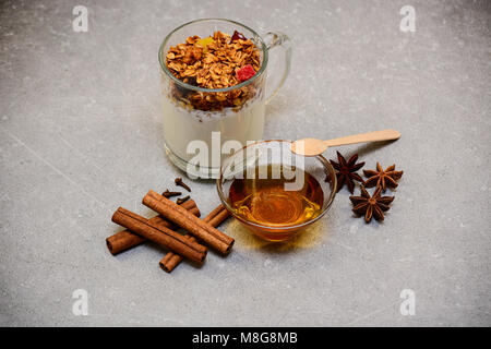 Müsli mit getrockneten Beeren, kandierten Früchten, Nüssen, Joghurt, goldenem Honig, Anis, Zimtstangen und Nelken auf steinernem Hintergrund. Stockfoto