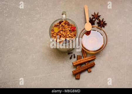 Müsli mit getrockneten Beeren, kandierten Früchten, Nüssen, Joghurt, goldenem Honig, Anis, Zimtstangen und Nelken auf steinernem Hintergrund. Stockfoto