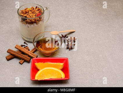 Granola mit getrockneten Beeren, kandierten Früchten, Nüssen, Joghurt, goldenem Honig, Orange, Anis, Zimtstangen und Nelken auf steinernem Hintergrund. Stockfoto