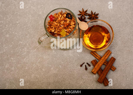Müsli mit getrockneten Beeren, kandierten Früchten, Nüssen, Joghurt, goldenem Honig, Anis, Zimtstangen und Nelken auf steinernem Hintergrund. Stockfoto