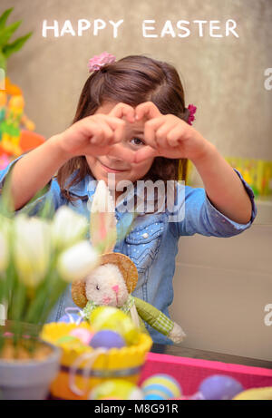 Süße kleine Mädchen vorbereiten Der frohe Ostern Urlaub mit Ihrem kleinen Freund Bunny Toy, zeigt Herz Geste, dekorieren Eier in verschiedenen Farben, Liebe Stockfoto