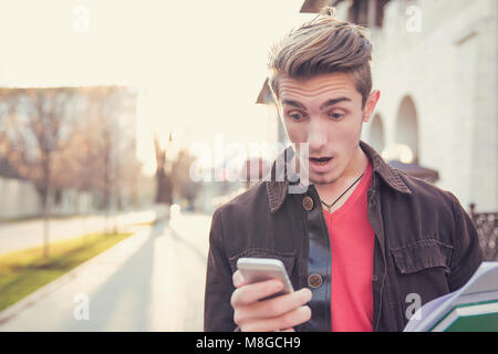 Junge legerer Mann mit Studien prüfen news Smartphone extrem auf städtischen Hintergrund geschockt. Stockfoto