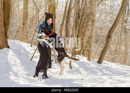 Stilvolle Frau mit lustigen Hut und Rock mit ihr spielen Siberian husky Hund im Winter Wald mit dem Stick Stockfoto
