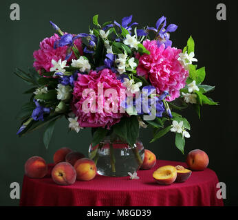 Blumenstrauß aus kultivierten Blumen in die Kanne und Pfirsiche auf einem runden Tisch mit einer roten Tischdecke. Stillleben mit Pfingstrose, Jasmin und Iris. Stockfoto