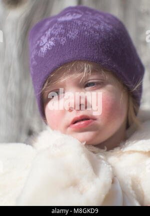 Candid outdoor winter Portrait von einem kleinen Mädchen mit blonden Haaren, blauen Augen und rosige Wangen, eine weisse Jacke und eine purpurrote Socke cap. Stockfoto