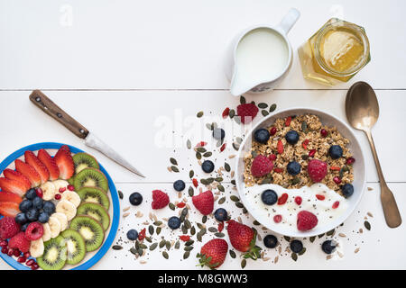 Frühstück Schüssel Müsli und einen Teller mit geschnittenem Obst mit Samen, Joghurt und Honig auf einem weißen Holz Hintergrund Stockfoto