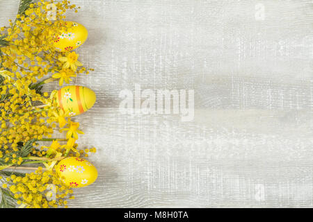 Zusammensetzung mit bemalten Eiern. Ostern Zubehör, Mimosa und gelbe Narzissen auf hellem Holz- Oberfläche. Gelb Orange Ostern Konzept, Ansicht von oben, kopieren Stockfoto