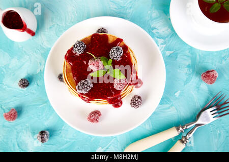 Amerikanische Pfannkuchen mit Marmelade und gefrorene Himbeeren mit Tee auf blauem Hintergrund. Ansicht von oben. Stockfoto