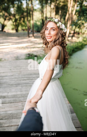 Die Braut im weißen Kleid ist die Hand des Bräutigams in einem Park in der Nähe von See. Folgen Sie mir Stockfoto
