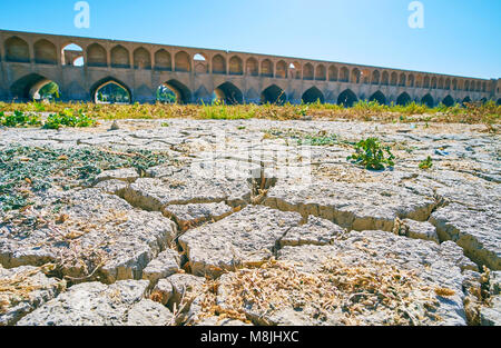 Der Spaziergang über die Ausgetrockneten Zayandeh Fluss, die Risse im Schlamm seiner Unterseite wird mit schlechter Vegetation, Si-o-se-pol Bridge auf dem Hintergrund zu sehen ist, Stockfoto