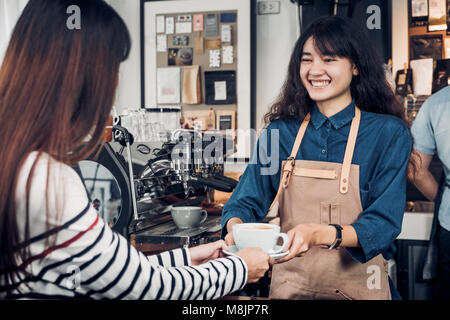 Asiatische Frau barista Schürze tragen Jean holding Tasse Kaffee serviert, um Kunden an Theke mit Smile Emotion, Cafe Restaurant Service Konzept, Inhaber smal Stockfoto