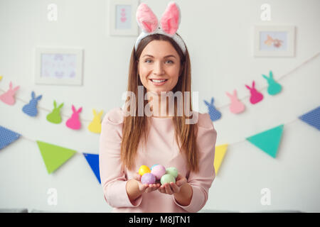 Junge Frau zu Hause Ostern celbration Konzept tragen Häschenohren Holding bunte Eier suchen Kamera lächelt Fröhlich Stockfoto