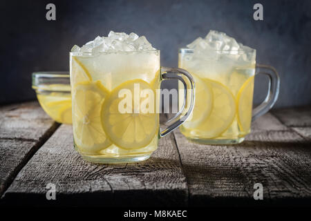 Zwei Tassen Limonade und Eiswürfel Krümel auf einem Holztisch Hintergrund natürlicher Zitrone Wasser essen Soda Drink mit Zitronenscheiben in eine Schüssel geben. Stockfoto