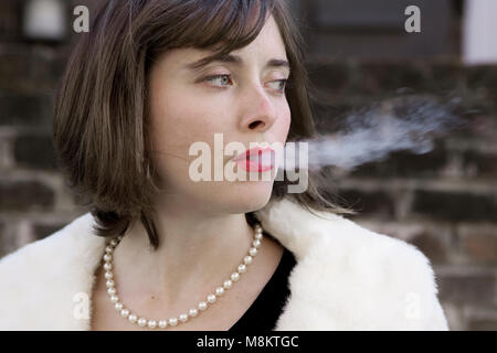 Eine gut gekleidete Frau das Tragen der roten Lippenstift und eine Perle Halskette, ausatmen Zigarettenrauch. Stockfoto