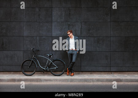 Stilvolle Junge Unternehmer in Sonnenbrille steht in der Nähe ein Fahrrad, mit dem Rücken gegen eine Wand brickgray Stockfoto