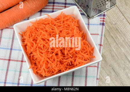 Konzept für die Vorbereitung einer gesunden Salat - geriebenen Karotten in einer weißen Platte auf einem Hintergrund von reibeisen und Karotten. Stockfoto