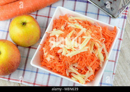 Konzept für die Vorbereitung einer gesunden Salat - geriebenen Karotten mit Apfel in einer weißen Platte auf einem Hintergrund von reibeisen, Karotten und Äpfel. Stockfoto