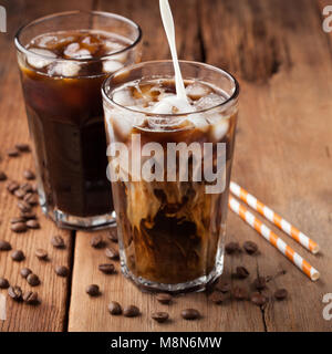 Eis Kaffee in ein hohes Glas mit Sahne über und Kaffeebohnen auf einem alten rustikalen Holztisch gegossen. Kalten Sommer trinken auf einem dunklen Hintergrund Stockfoto