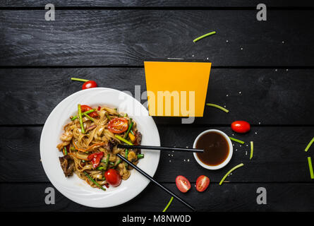 Udon fry Nudeln mit Meeresfrüchten und Champignons in eine weiße Platte auf schwarzem Hintergrund rühren. Mit Stäbchen und Box für Nudeln. Stockfoto