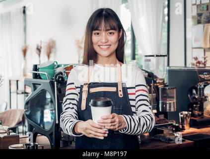 Asiatische Frau barista Schürze tragen Jean holding Hot nehmen Sie Kaffee Tasse bedient Kunden mit lächelnden Gesicht an der Theke. Cafe Restaurant Service conce Stockfoto