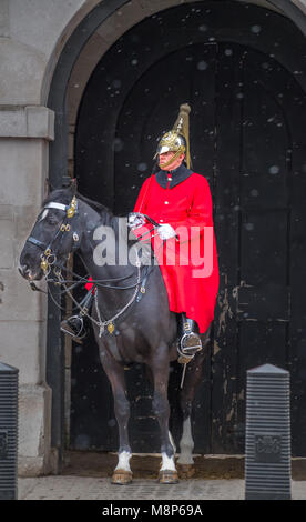 Kavallerie Soldat, der mit seinem metallischen Helm und in seinem roten Mantel, auf einem Pferd an der Whitehall Tor der Horseguards Parade an einem verschneiten Frühling montiert Ein Stockfoto