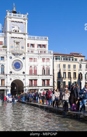 Touristen zu Fuß auf passerelles Vor dem Uhrturm durch eine überflutete Markusplatz, Piazza San Marco, Venedig, Italien während der Acqua Alta hig Stockfoto