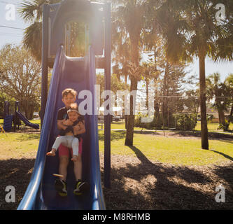 Bruder mit Schwester gleiten auf Folie am Spielplatz während der sonnigen Tag Stockfoto