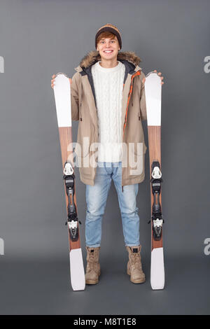 Junger Mann tragen Winterkleidung Studio auf graue Wand suchen Kamera lächelt Fröhlich holding Skier mit vollem Körper geschossen isoliert Stockfoto