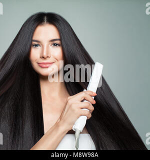 Friseursalons. Frau mit schönen langen glattes Haar mit Haarglätter. Süß lächelnde Mädchen richten Gesund Brünette Haare mit Bügeleisen. Uhr Stockfoto