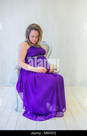 Schöne schwangere Frau in Ultra Violet Kleid sitzt auf dem Sessel Stockfoto