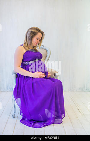 Schöne schwangere Frau in Ultra Violet Kleid sitzt auf dem Sessel Stockfoto
