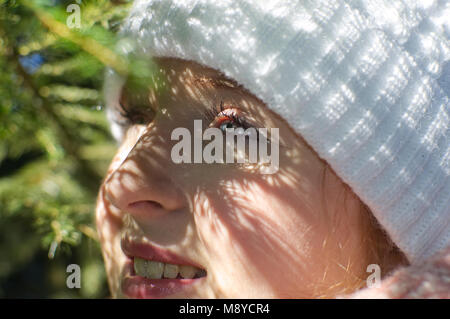 Eine junge Frau mit einem weißen Hut und blauen Augen hinter Zweigen. Schatten und Licht. Stockfoto