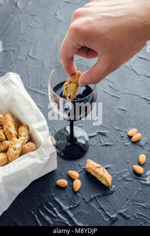Männliche hand Tauchen traditionelle italienische Almond cookies Cantuccini in Rotwein auf dem schwarzen Stein. Hausgemachten biscotti auf dunklem Beton Tabelle. Stockfoto