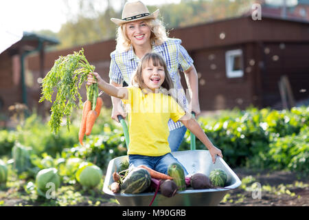 Frau schob ihren Kind Tochter in einer Schubkarre gefüllt Gemüse im Garten Stockfoto