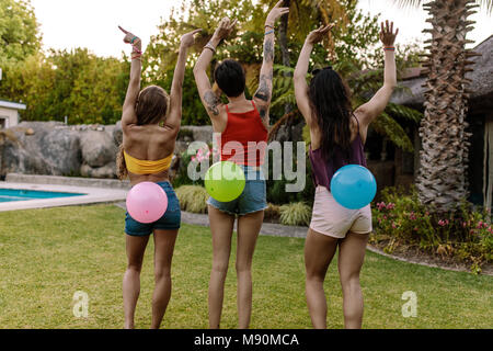 Ansicht der Rückseite drei Frauen stehen im Freien mit Ballons auf dem Rücken gebunden. Freunde spielen Ballon platzen Spiel auf einer Party. Stockfoto