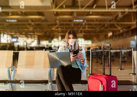 Flughafen jungen weiblichen Passagier auf Smartphone und Laptop im Terminal Hall beim Warten auf ihren Flug Stockfoto