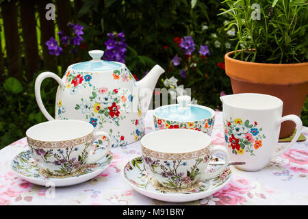 Ein blumiger Kaffee Service mit Teekanne, Tassen und Untertassen und Milchkännchen an einem Picknicktisch in einem englischen Country Garden bereit für Tee am Nachmittag. Stockfoto