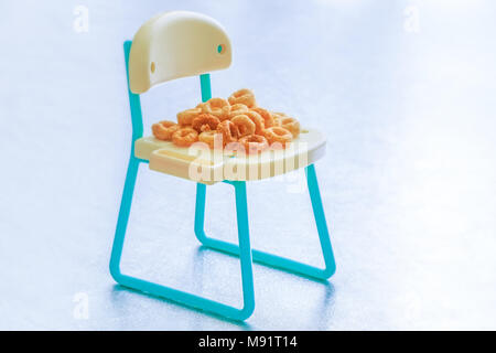 Stapel der eine Menge runde Frühstückszerealien auf miniatur Stuhl isoliert. Gesundes Müsli Ringe auf dem Sitz. Konzeptionelle Bild für Getreide, Kinder essen. Stockfoto