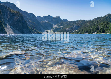 Unberührten alpinen See im Sommer von immergrünen Pinienwald Mitte tag Sun close up Low Angle Wasser umgeben. Stockfoto