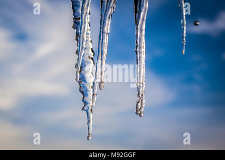 Große Eiszapfen auf einem Hausdach, blauer Himmel und Wolken im Hintergrund. Eiszapfen hängen vom Dach eines Hauses, winter Thema. Stockfoto